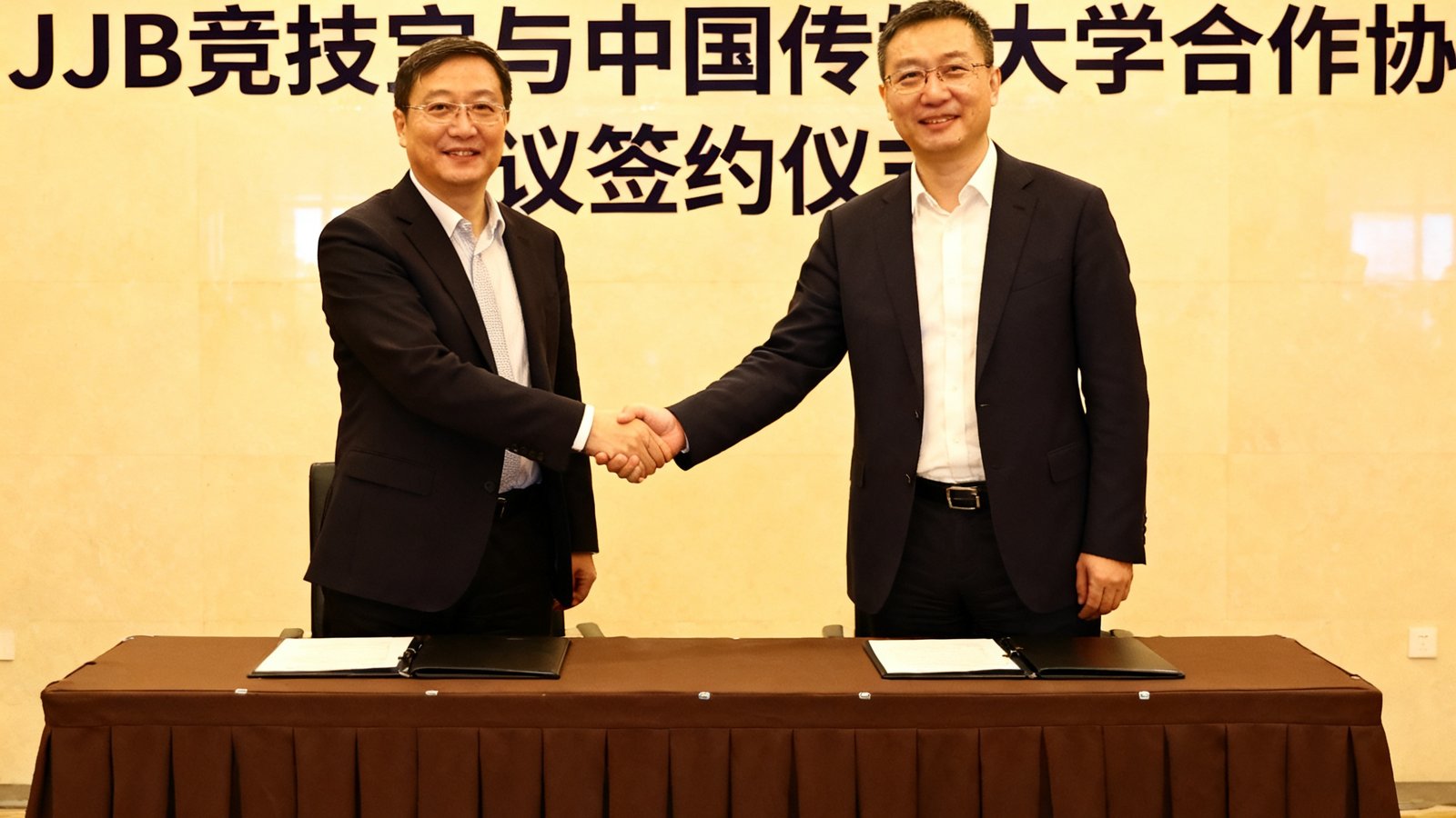 JJB竞技宝与中国传媒大学签署合作协议的现场照片，双方代表在签约台前握手，背景墙上印有双方Logo和合作标题。